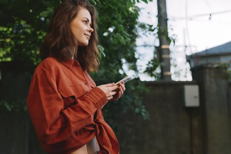 Young woman with wavy hair wearing an orange shirt checks her smartphone outdoors, surrounded by lush greenery, creating a casual lifestyle moment with warm natural light and a relaxed mood