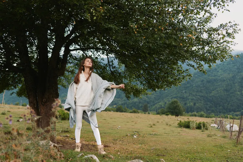 Casual lifestyle portrait of a woman standing in a grassy field with trees in the background, relaxed pose, soft natural light, open landscape, serene outdoor mood
