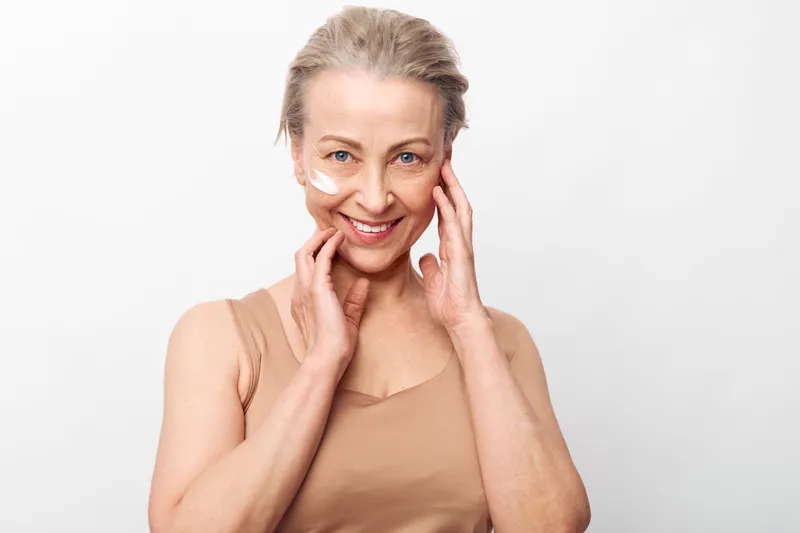 Smiling mature woman with natural skin applies cream on cheeks, showing skincare routine and self-care. Close-up portrait with light beige clothing and bright white background for wellness concept.