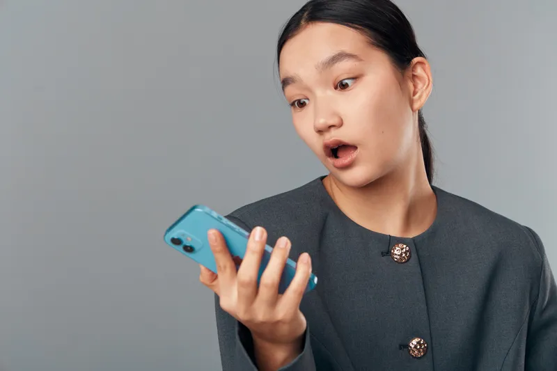 Young woman looking surprised at smartphone with open mouth, wearing gray jacket on gray background. Female person holding mobile device with shocked facial expression and raised eyebrows.