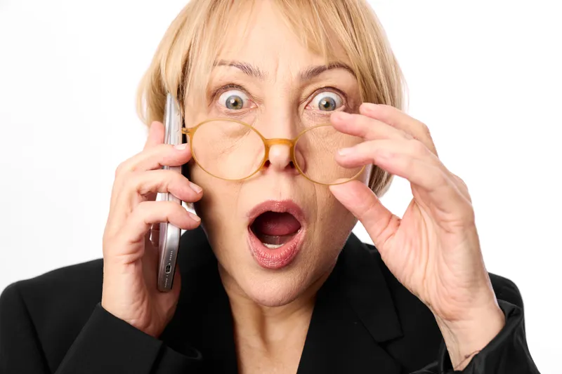 Surprised blonde woman holding glasses and a phone to her ear, close up, white studio background, amazed expression, concept of communication, eyewear, visual attention, style