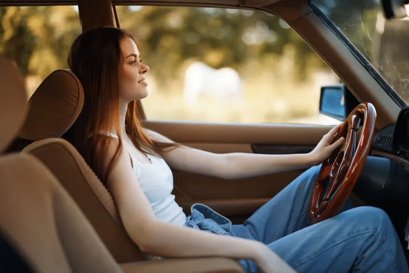 Young woman with long hair in casual outfit sits confidently in vintage car, enjoying summer vibes in natural background.