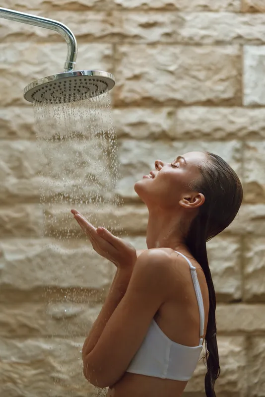 woman showering under outdoor rain showerhead, enjoying water stream, natural light, relaxed mood, stone wall background, wellness moment
