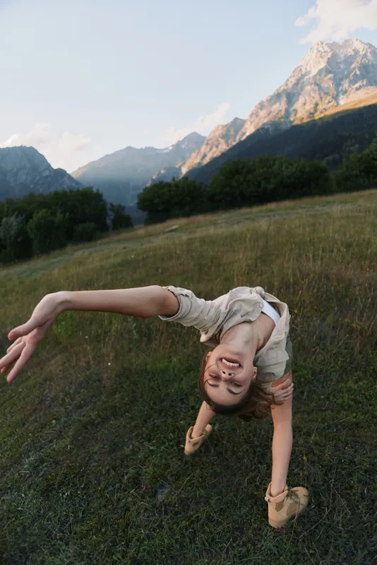 Young woman flips gracefully in a sunlit grassy field with distant mountains, expressing carefree playfulness and balance.