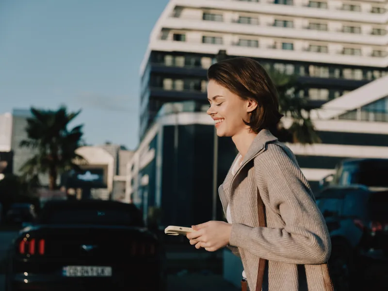A cheerful business professional walks outdoors, texting on a smartphone. She smiles warmly, radiating confidence and a positive, dynamic energy in a modern urban setting.