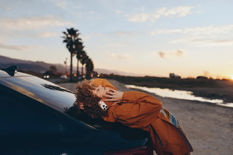 Woman lifestyle at sunset beachside, curly hair and orange jacket leaning on a parked car hood, relaxed hand on hair, warm golden light, travel vacation mood with blurred ocean waves in soft