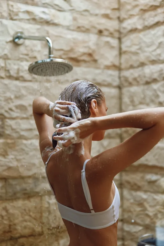 Woman washing hair under shower with soap foam, bare back visible, natural stone wall background, relaxed morning routine, personal care in warm lighting.
