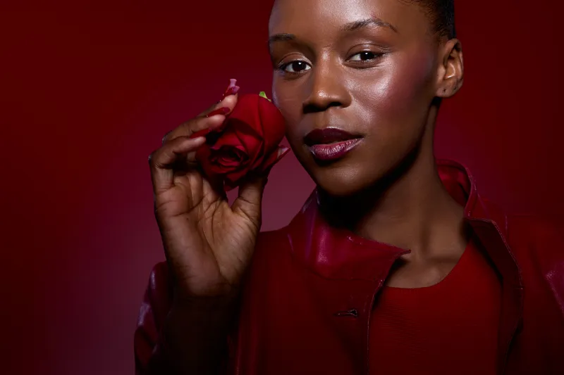 Close-up of a dark skinned African woman holding a red rose near her face. She wears a red leather jacket against a red background. Concept of beauty, elegance, and passion.