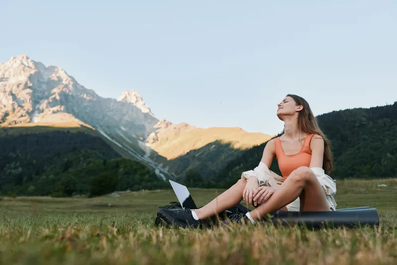 A cheerful woman sits on a grassy field with a laptop, looking up toward sunlit mountains. She appears focused and relaxed, enjoying a productive outdoor workspace.