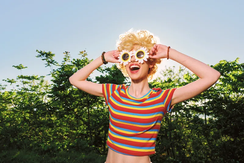 Cheerful woman outdoors, smiling with playful pose in a striped top, retro color palette, sunlit scene, lush greenery, carefree mood, candid lifestyle moment and sunny breeze adds