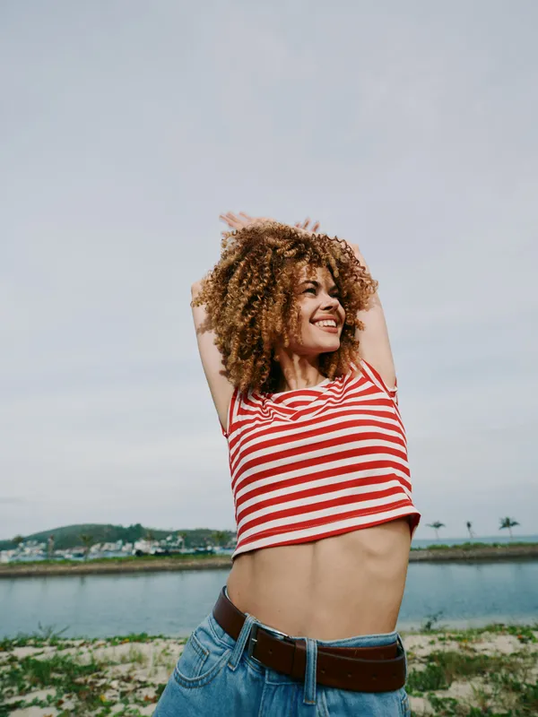 Young woman with curly hair in a red striped top and denim jeans stretches her arms above her head, smiling confidently near a calm waterfront under a bright sky.