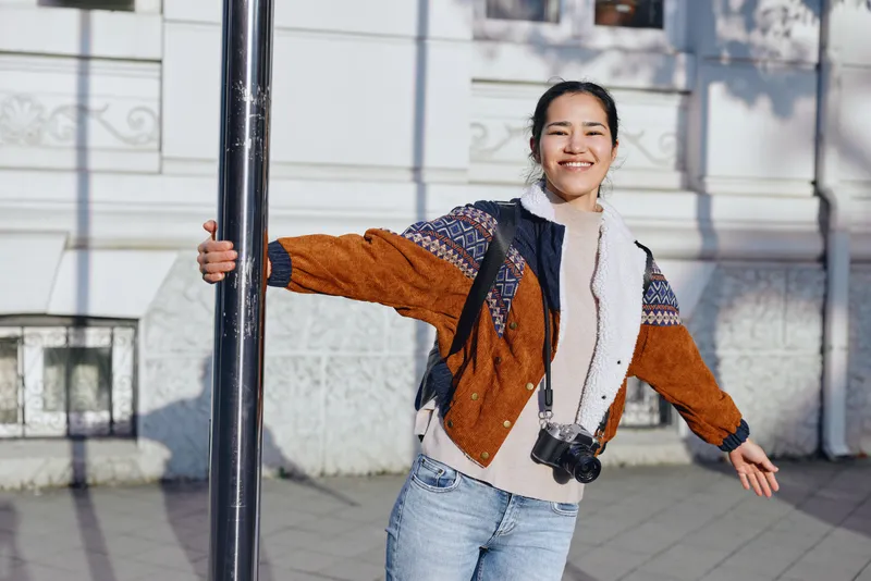 Joyful woman in denim jeans and warm jacket with scarf poses beside a street pole in an urban outdoor setting, smiling, embracing casual fashion and sunshine on a crisp day