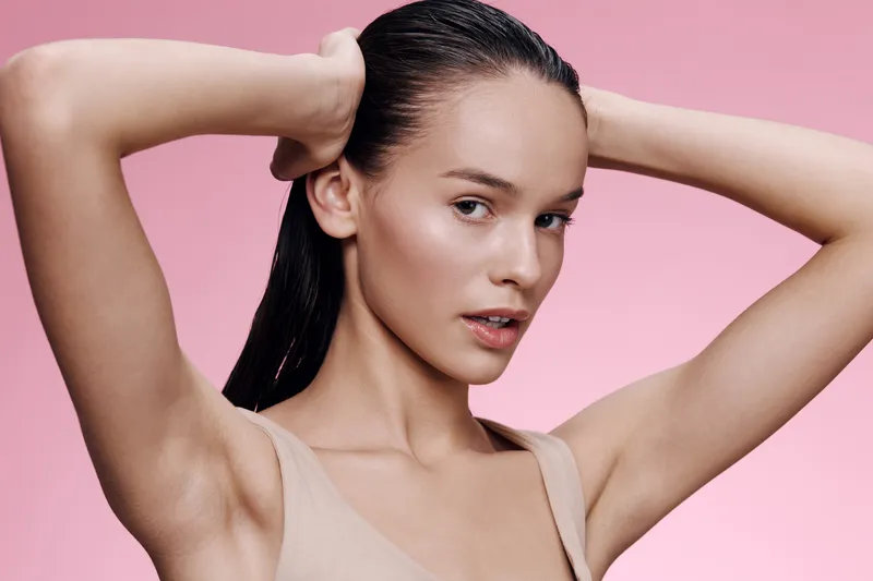 Young woman with wet hair in a beige top against a pink background, exuding confidence and elegance, showcasing beauty and modern aesthetics