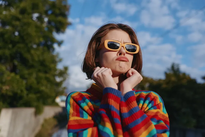 Young woman in early twenties wearing a colorful striped sweater and yellow sunglasses, playful pout expression and hands at chin in outdoor portrait, casual fashion lifestyle in a suburban park.