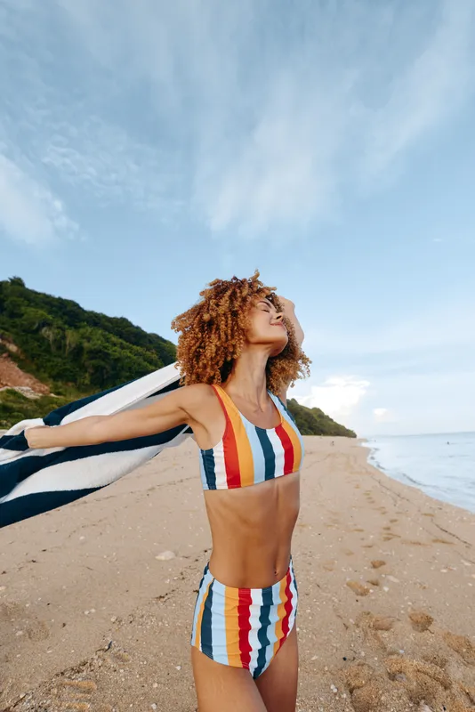 Young woman on a sunny beach wears a striped bikini and holds a surfboard, enjoying the ocean breeze, bright sky, and active summer mood along the sandy shoreline
