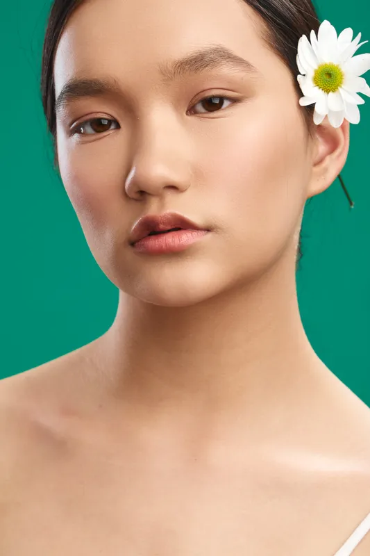 Asian woman with a white daisy tucked behind the ear stands against a vibrant green backdrop; she gazes softly, exuding calm, natural beauty in a studio portrait.