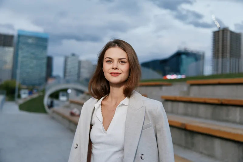 Confident businesswoman in a stylish suit, smiling outdoors with a modern cityscape background, showcasing professionalism and ambition.