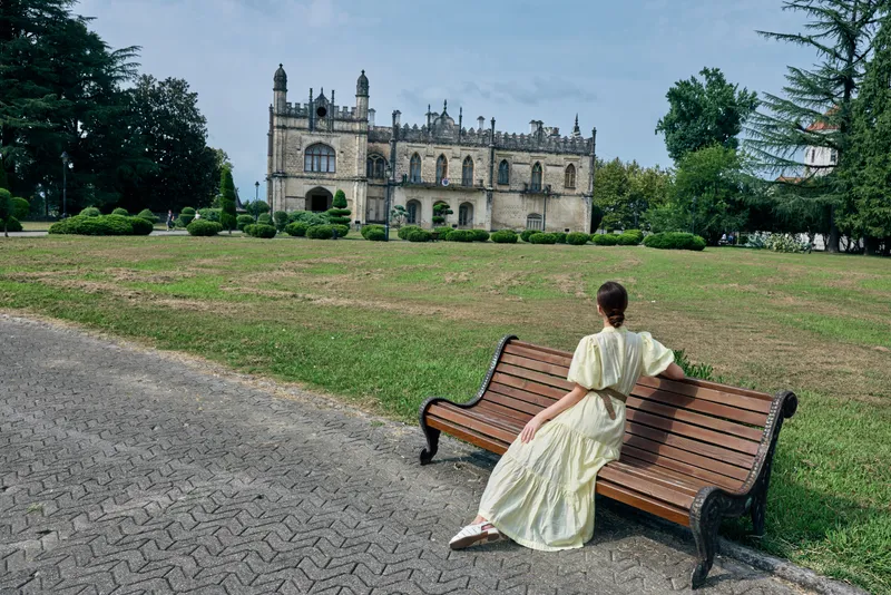 A woman in a flowing dress sits on a wooden bench in a grassy park, facing a historic castle, creating a calm outdoor travel moment and nostalgic scenic atmosphere.