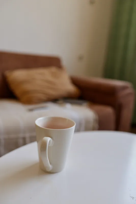 White mug on a round table in a cozy living room, soft natural light, calm morning mood, simple domestic scene, inviting everyday ritual and quiet comfort.