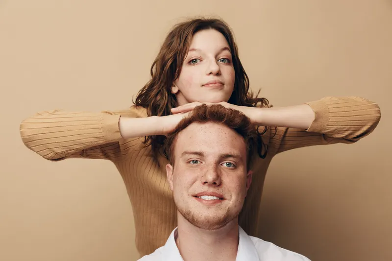 Young man and woman pose together on beige background. Woman rests hands on man’s head, both smiling gently. Concept of friendship, casual style, and relaxed mood in studio.