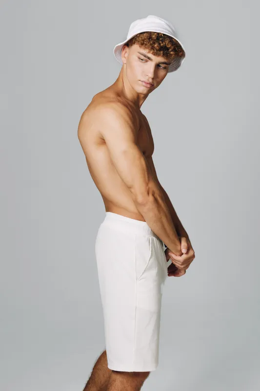 Young man with curly hair wearing white shorts and bucket hat posing shirtless against gray background, showing side profile and relaxed expression in studio lighting.