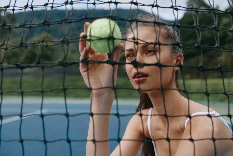 Young female tennis player holding a tennis ball, looking through a net on an outdoor court, exuding focus and determination during a sunny day