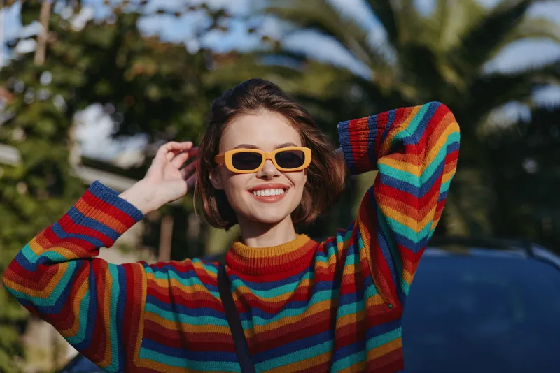 Young woman in mid twenties wearing a colorful rainbow sweater and yellow sunglasses smiling outdoors by palm trees. Short brown hair, casual outfit and joyful pose for lifestyle travel.