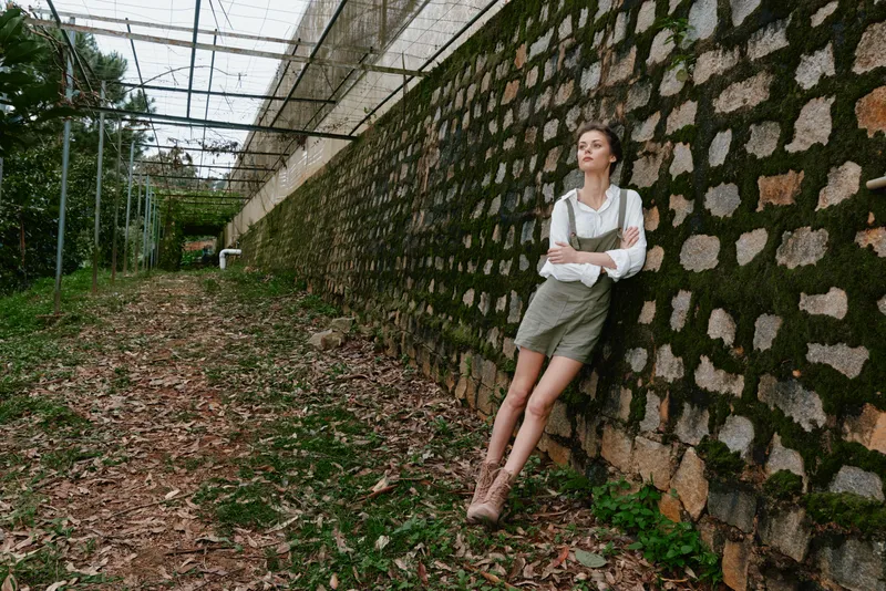 Young woman in casual summer outfit against rustic wall, showcasing relaxed emotions and natural beauty amidst greenery
