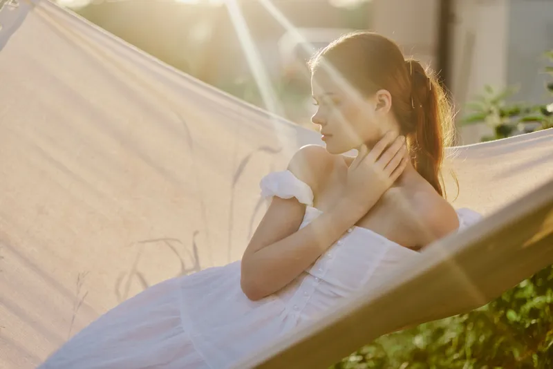 A serene young woman relaxing in a hammock, wearing a white dress, basking in sunlight and greenery, embodying tranquility and summer vibes
