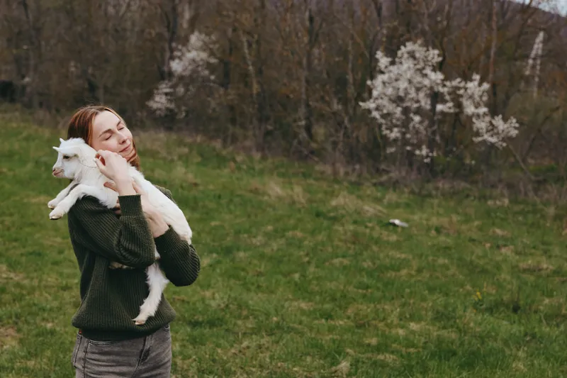 Woman outdoors in a green field holding a goat kid, showing gentle bonding and rustic lifestyle. Natural light and open countryside convey joy, tenderness, and a strong connection with animals.