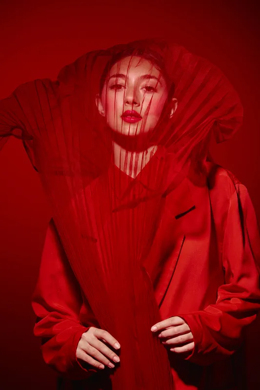 Woman in a bright red fashion coat poses with dramatic red concealer and mesh fabric covering her face. Creative beauty shot with bold color styling and avant-garde concept.