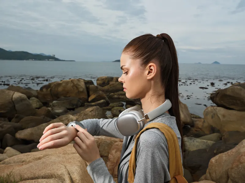 Young woman checking time on smartwatch by the rocky beach, wearing headphones, casual outfit, serene emotions, coastal background, modern lifestyle concept