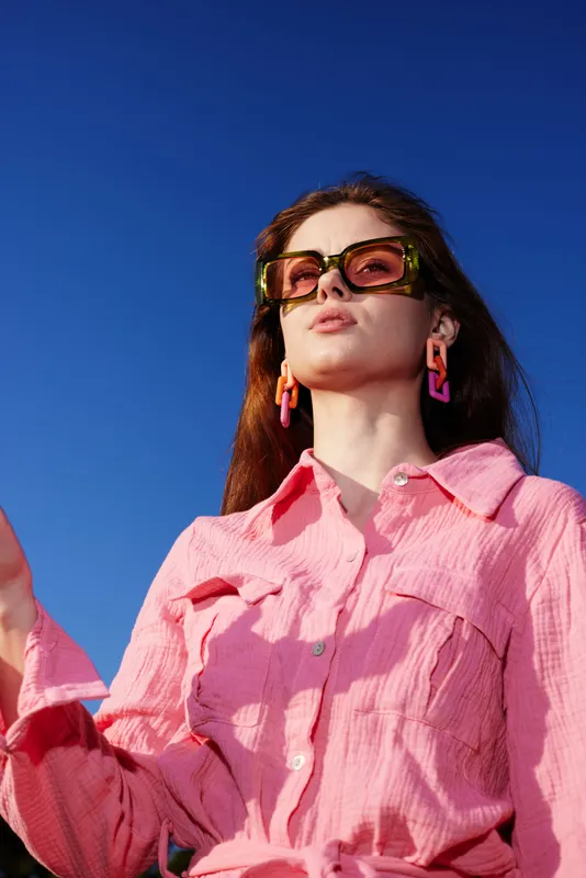 Young woman with brown hair wearing a pink shirt and stylish sunglasses against a blue sky backdrop, exuding confidence and summer vibes, ideal for fashion and lifestyle imagery