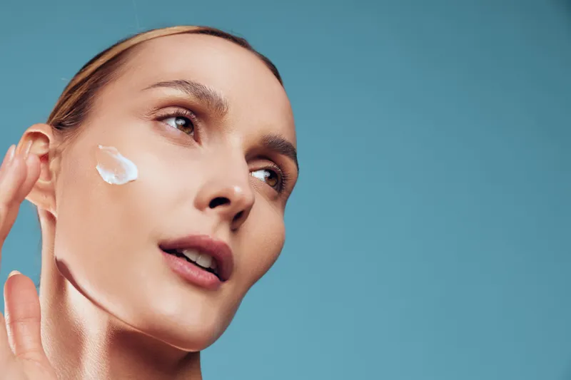 Young woman with brunette hair applies white cream to her cheek in a close-up studio shot, highlighting skincare, wellness, and a calm confident mood