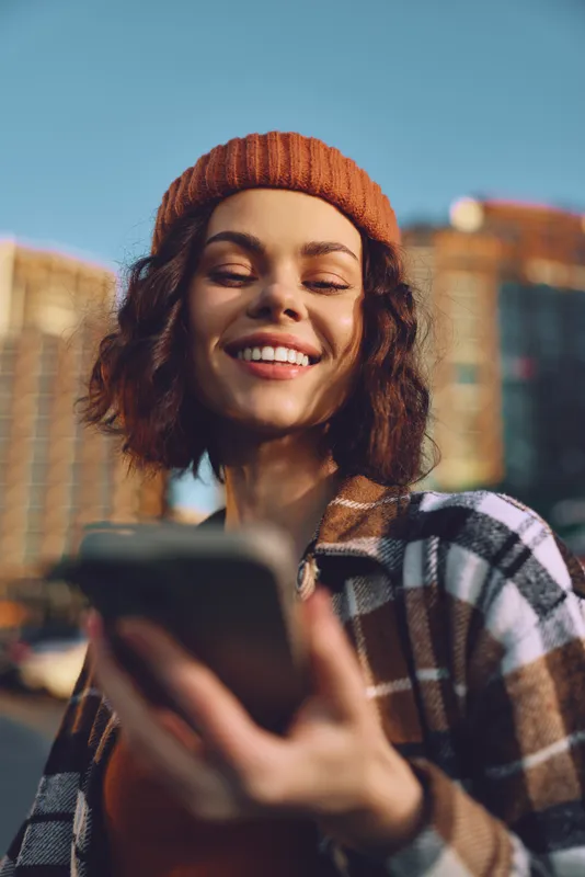 Woman smiling with phone in urban street during golden hour glow, beanie and plaid jacket conveying authenticity and candid lifestyle, mindful living and emotional storytelling.