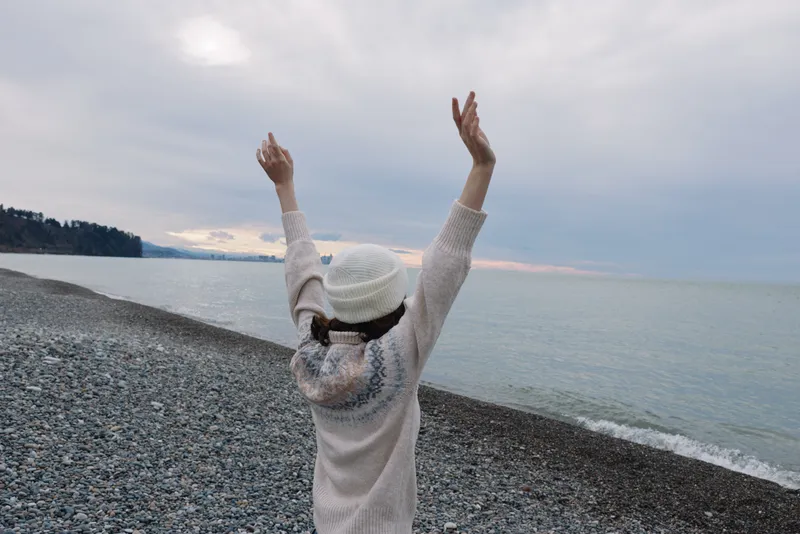 Beach scene with a person facing the sea, arms raised in celebration, white hat and sweater, pebble shoreline, vast ocean horizon, peaceful outdoor moment of joy and freedom