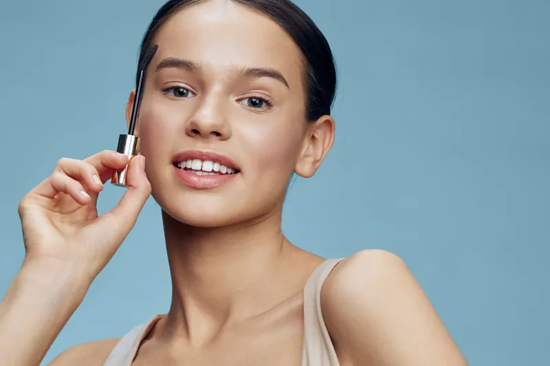 Young woman applying cosmetic product with a joyful smile, showcasing healthy skin against a soft blue background, ideal for beauty and skincare concepts