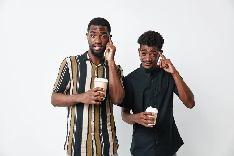 Two young African men with dark skin holding coffee cups and pointing to their heads on white background. Casual style, friendship and thoughtful gesture concept.