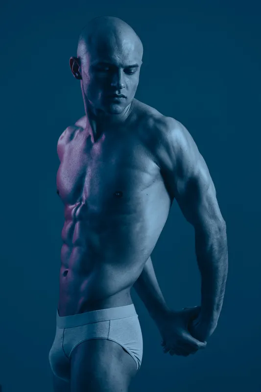 Strong man posing with a focused expression, showcasing his muscular physique in a dramatic blue toned setting. This image captures strength and determination beautifully.