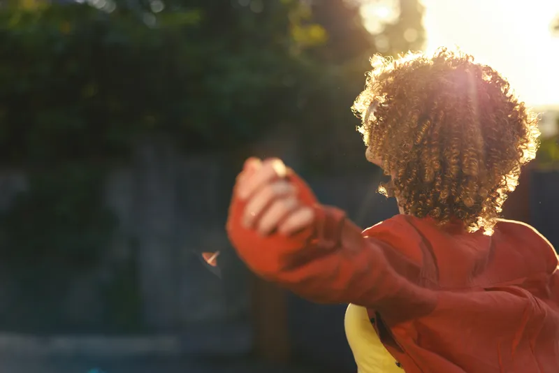 Bright candid shot of a woman outdoors, back facing camera, curly hair, red jacket, arms raised in joyful motion, warm sunlight, relaxed lifestyle moment in a park with soft bokeh, everyday happiness