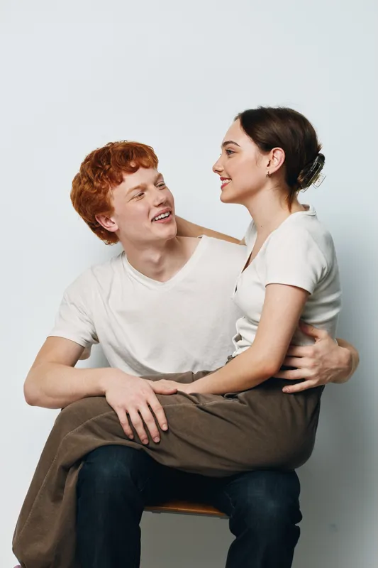 Couple, young man and woman sitting together in casual clothing smiling and embracing on white background. Relationship, affection and friendship with happiness and comfort indoors.
