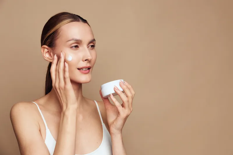 Young woman with brunette hair applies cream on her face while holding a white jar against a beige background in a skincare beauty routine.
