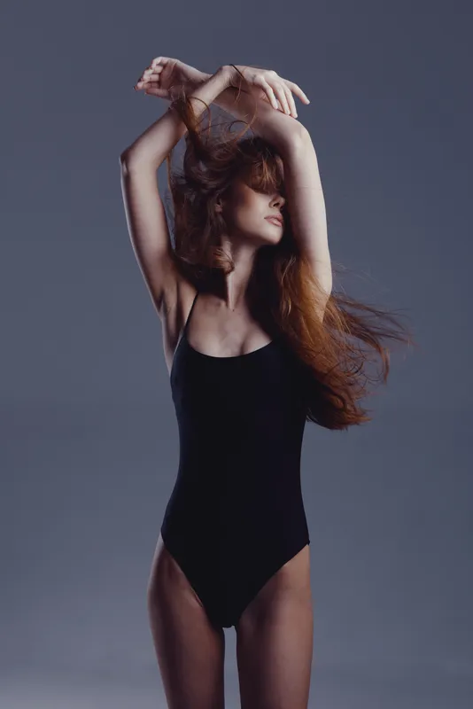 Young woman in black swimsuit posing with raised arms against gray background, showcasing elegance and summer style.