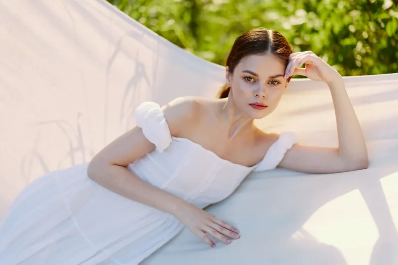 Elegant young woman in a white dress poses gracefully in a serene outdoor setting, embodying beauty, tranquility, and femininity
