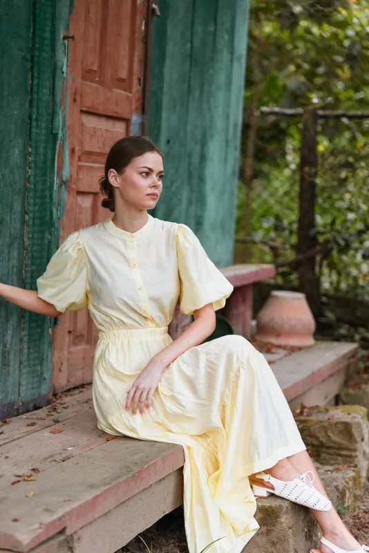 Woman sits on steps in a pale yellow dress by a rustic doorway, relaxed pose and soft natural light creating a calm outdoor mood, her profile reveals poise and quiet confidence