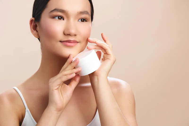 asian woman applies moisturizer in a soft light setting, smiling gently as she cares for her skin, presenting a calm and confident beauty routine in a minimal studio.