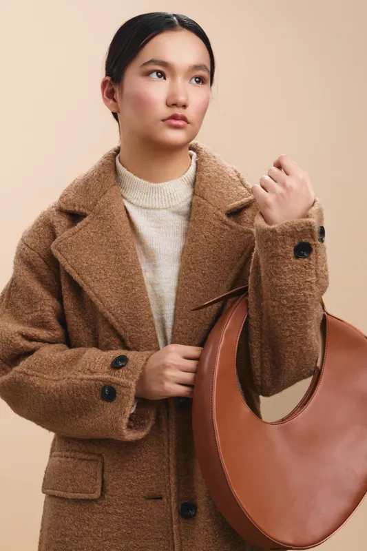 Asian woman wearing a tan wool coat poses with a brown handbag against a neutral backdrop, presenting a stylish, composed look that exudes confidence and refined fashion.