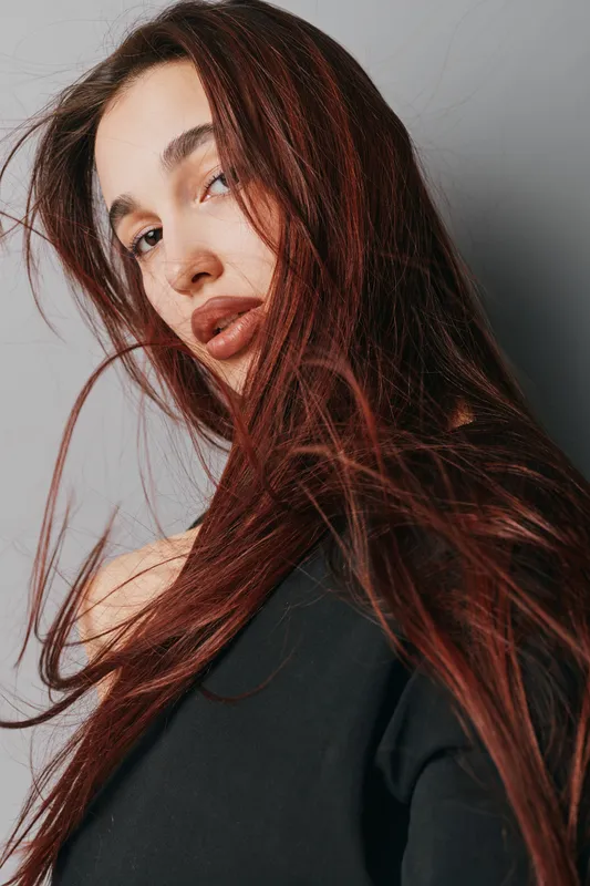 Woman with long red hair and natural makeup posing against gray background, wearing black top with loose strands framing her face in soft light portrait photography.
