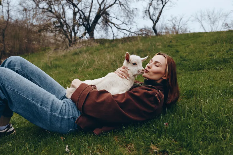 Joyful woman outdoors, relaxing on grass with a goat kid, gentle cuddle, sunlit moment, casual lifestyle scene, natural textures, cozy attire, green hillside, peaceful companionship, carefree mood