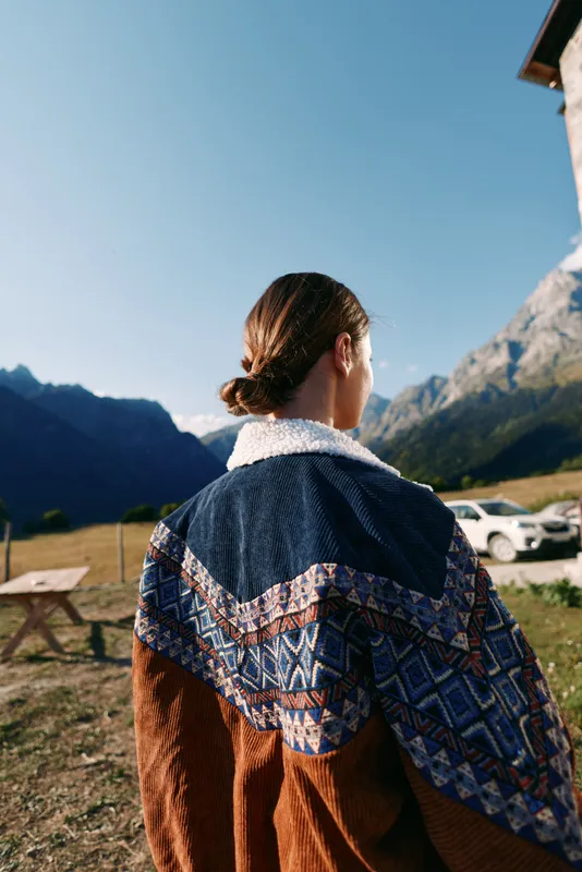 Woman in a patterned sweater stands with her back to camera in the mountains outdoors, overlooking a quiet countryside landscape under clear sky, enjoying autumn light and peaceful nature.